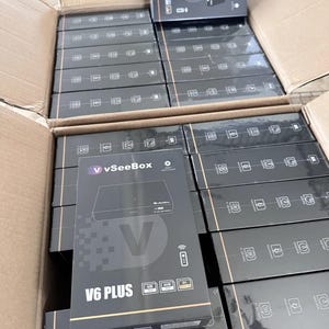 May include: A cardboard box filled with multiple black V6 PLUS vSeeBox media player boxes. Each box features the vSeeBox logo, product name, and specifications. The boxes are stacked neatly inside the open cardboard container, ready for distribution.
