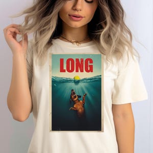 May include: Cream-colored t-shirt featuring a graphic print. The print depicts a dachshund dog underwater, reaching for a yellow ball. The word "LONG" is printed in red above the dog.