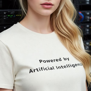 Puede incluir: Una mujer rubia con una camiseta crema con el texto "Powered by Artificial Intelligence" en negro. Está de pie frente a un rack de servidores con cables.