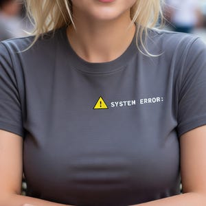 May include: A gray t-shirt with the text "SYSTEM ERROR:" and a yellow warning symbol. A white coffee cup and saucer sit on a round table. The person is sitting outside at a cafe.