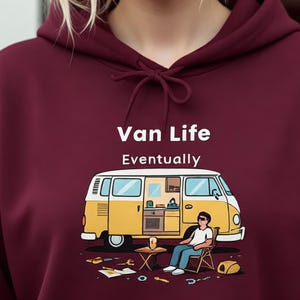 Pode incluir: Um moletom bordô com o texto "Van Life Eventually" e uma ilustração de uma van amarela. A ilustração mostra uma pessoa relaxando fora da van. A pessoa na imagem está sorrindo.