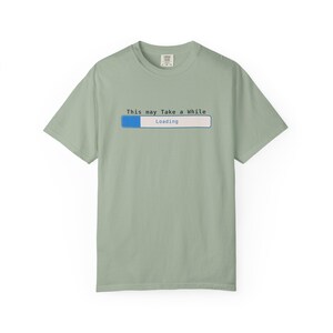 Puede incluir: Camiseta verde salvia con el texto "This may Take a While" sobre un gráfico de barra de carga azul y blanco. La palabra "Loading" está debajo de la barra. La camiseta tiene mangas cortas y cuello redondo.