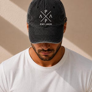 Könnte beinhalten: Eine schwarze, ausgewaschene Baseballkappe mit gebogenem Schirm. Die Kappe zeigt ein weißes Grafikdesign mit den Buchstaben "P" und "A" und dem Text "EST. 2026". Sie hat einen verstellbaren Riemen.