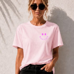 Pode incluir: Uma t-shirt de manga curta cor-de-rosa claro com um pequeno gráfico roxo no peito esquerdo. O gráfico consiste em dois círculos roxos sólidos para os olhos e a palavra WHATEVER escrita num arco roxo por baixo para formar um rosto.
