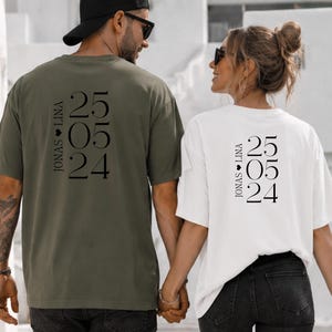 Può includere: Una t-shirt verde oliva e una t-shirt bianca, entrambe con testo nero sul retro che riporta JONAS CUORE LINA e la data 25 05 24 in verticale. Le t-shirt hanno maniche corte e scollo rotondo, indossate con pantaloni di colore nero.