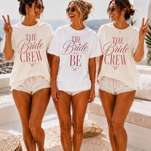 May include: Three short-sleeved crewneck t-shirts. Two off-white shirts feature the red text THE Bride CREW EST. 2026 with a small bow icon. The center white shirt features the red text THE Bride TO BE EST. 2026 with a small bow icon.