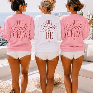 May include: Two pink sweatshirts with the text THE BRIDE CREW EST. 2026 and one white sweatshirt with the text THE BRIDE TO BE EST. 2026 in dark red lettering. Each sweatshirt features a small bow graphic below the text and long sleeves.