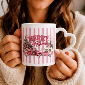 Op de afbeelding: Een witte keramische mok met een hartvormig handvat en een verticaal gestreept patroon in roze en wit. Het ontwerp toont een roze pick-up truck die een roze caravan trekt, versierd met kerstbomen, cadeautjes en de tekst Merry Christmas.