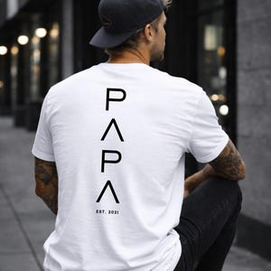 May include: White t-shirt with the word "PAPA" in black capital letters stacked vertically on the back, with "EST. 2021" below. The person is wearing a dark gray baseball cap and black jeans.