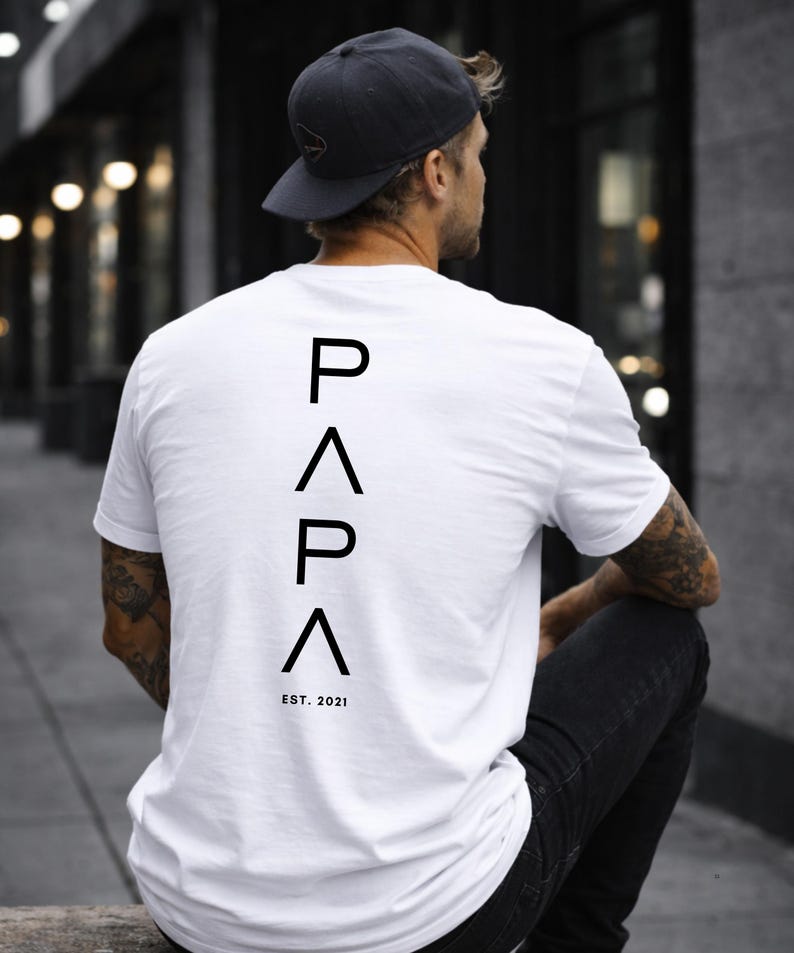 May include: White t-shirt with the word "PAPA" in black capital letters stacked vertically on the back, with "EST. 2021" below. The person is wearing a dark gray baseball cap and black jeans.