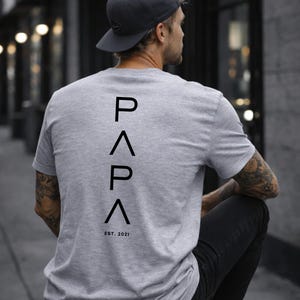 May include: Gray t-shirt with the word "PAPA" in black, with an upward-pointing triangle above and below the letters, and the text "EST. 2021" below. The person is wearing a gray cap and black pants.