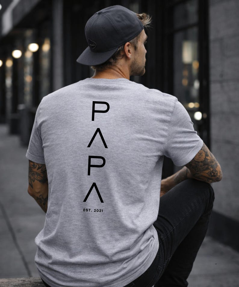 May include: Gray t-shirt with the word "PAPA" in black, with an upward-pointing triangle above and below the letters, and the text "EST. 2021" below. The person is wearing a gray cap and black pants.