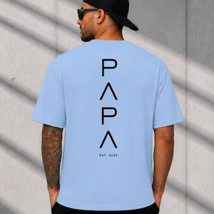 May include: Light blue t-shirt with the word "PAPA" in black, stacked vertically, and the text "EST. 2025" below. The shirt is short-sleeved and has a crew neck. The person is wearing a black cap and sunglasses.