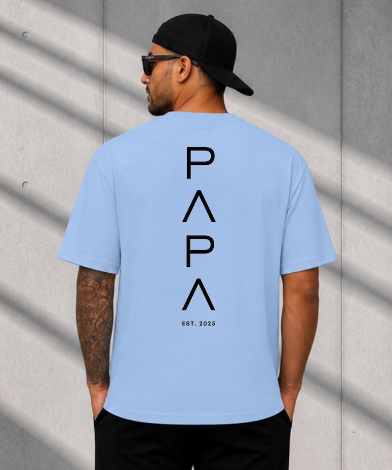May include: Light blue t-shirt with the word "PAPA" in black, stacked vertically, and the text "EST. 2025" below. The shirt is short-sleeved and has a crew neck. The person is wearing a black cap and sunglasses.