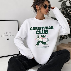 Könnte beinhalten: Ein weißes Sweatshirt mit Rundhalsausschnitt und einem dunkelgrünen Aufdruck mit der Aufschrift CHRISTMAS CLUB. Das Design zeigt eine Illustration einer Person mit Geschenken, den Text LET'S GET COZY und MERRY DAYS, COZY NIGHTS & CHRISTMAS VIBES.