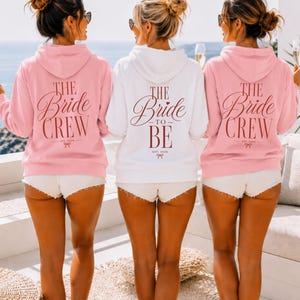 May include: Two light pink hoodies and one white hoodie featuring the text THE BRIDE CREW or THE BRIDE TO BE in dark red script lettering on the back. Each hoodie includes the text EST. 2026 and a small bow graphic below the main lettering.