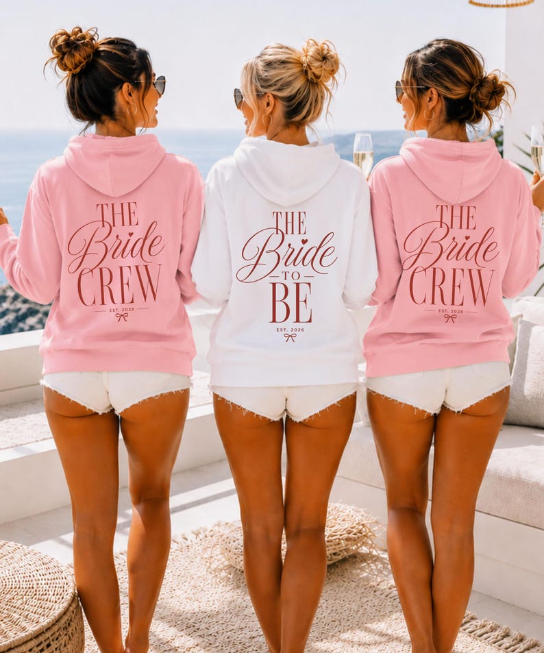 May include: Two light pink hoodies and one white hoodie featuring the text THE BRIDE CREW or THE BRIDE TO BE in dark red script lettering on the back. Each hoodie includes the text EST. 2026 and a small bow graphic below the main lettering.