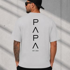 May include: A light gray t-shirt with the word "PAPA" stacked vertically in black, with an upward-pointing triangle between the letters. The text "EST. 2025" is below the word "PAPA".
