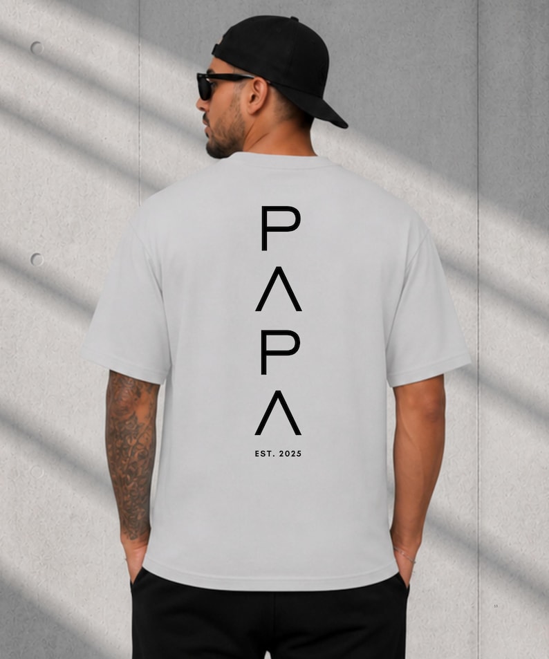 May include: A light gray t-shirt with the word "PAPA" stacked vertically in black, with an upward-pointing triangle between the letters. The text "EST. 2025" is below the word "PAPA".