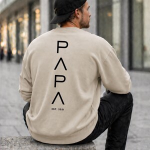 May include: Beige sweatshirt with the word "PAPA" printed vertically in black capital letters on the back. Below the word "PAPA" is the text "EST. 2021". The person in the image is wearing a black baseball cap and black pants.