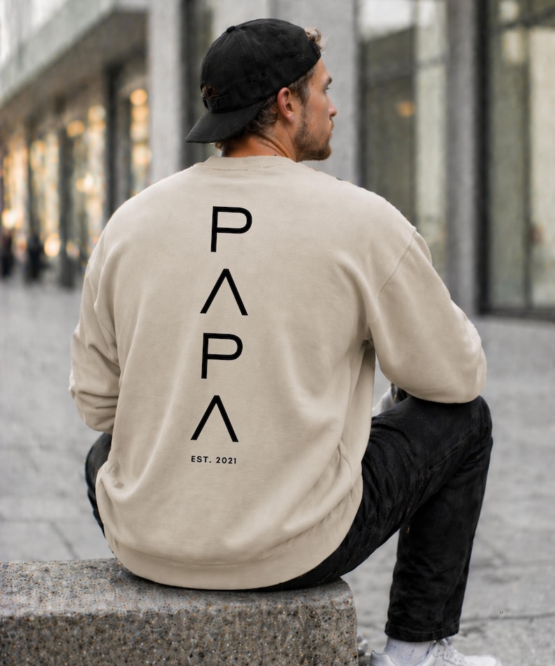 May include: Beige sweatshirt with the word "PAPA" printed vertically in black capital letters on the back. Below the word "PAPA" is the text "EST. 2021". The person in the image is wearing a black baseball cap and black pants.