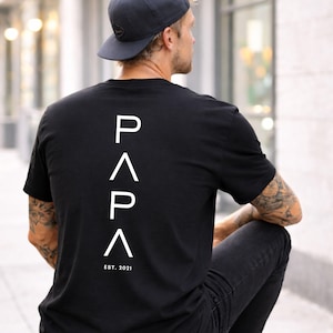 May include: Black t-shirt with the word "PAPA" in white, vertically aligned on the back, with "EST. 2021" below. The person in the image is wearing a dark blue baseball cap and black jeans.