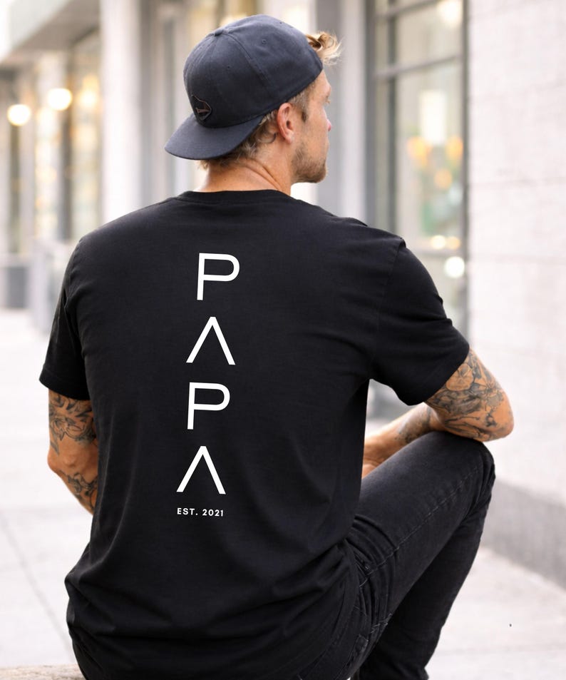 May include: Black t-shirt with the word "PAPA" in white, vertically aligned on the back, with "EST. 2021" below. The person in the image is wearing a dark blue baseball cap and black jeans.