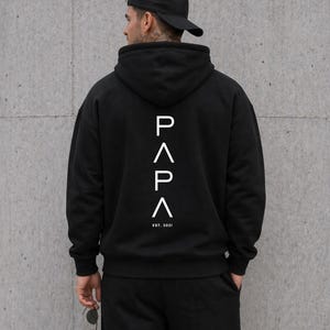 May include: Black hoodie with the word "PAPA" in white, with an upward-pointing arrow above and below the letters. The text "EST. 2021" is below the word "PAPA". The person in the image is wearing a black baseball cap and black pants.