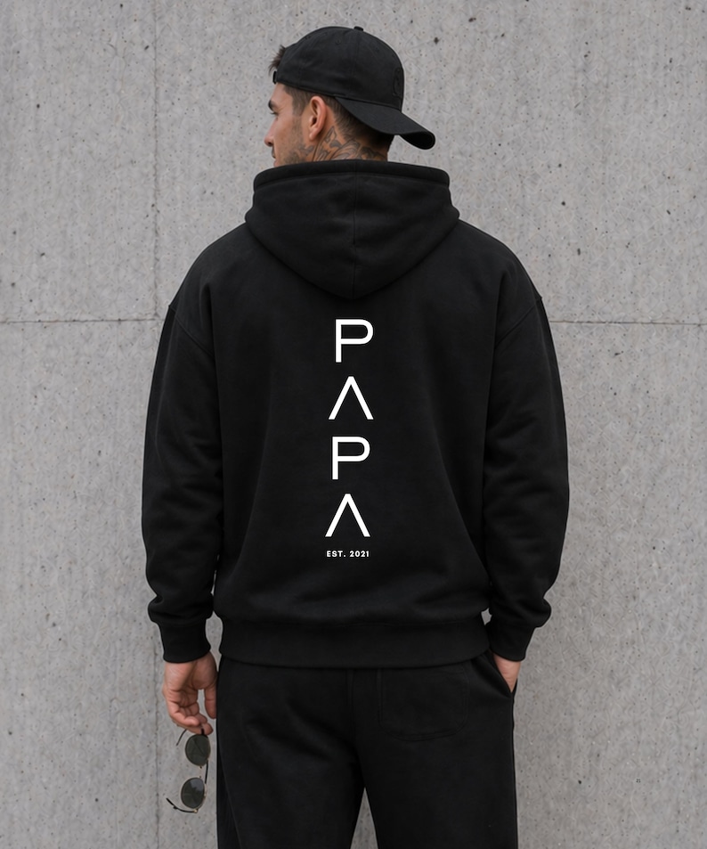May include: Black hoodie with the word "PAPA" in white, with an upward-pointing arrow above and below the letters. The text "EST. 2021" is below the word "PAPA". The person in the image is wearing a black baseball cap and black pants.