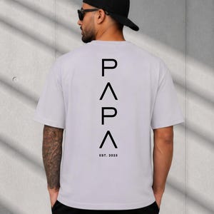 May include: A light gray t-shirt with the word "PAPA" in black, with an inverted triangle between the letters. Below the word is "EST. 2025". The shirt is short-sleeved.