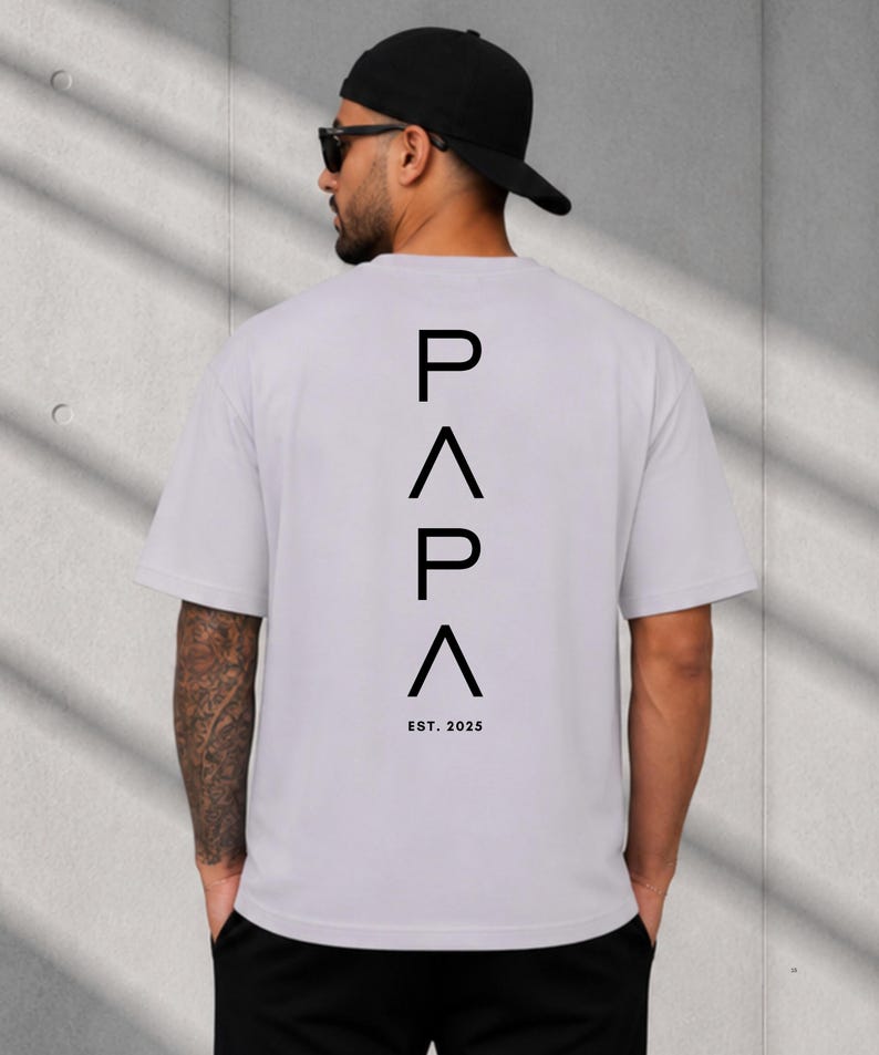 May include: A light gray t-shirt with the word "PAPA" in black, with an inverted triangle between the letters. Below the word is "EST. 2025". The shirt is short-sleeved.