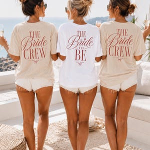 May include: Two beige t-shirts and one white t-shirt with short sleeves. The beige shirts feature the red text THE Bride CREW EST. 2024 with a bow icon. The white shirt features the red text THE Bride TO BE EST. 2024 with a bow icon.