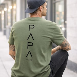 May include: Olive green t-shirt with the word "PAPA" printed vertically in black, with "EST. 2021" below. The shirt is worn by a person wearing a dark gray baseball cap and black jeans.