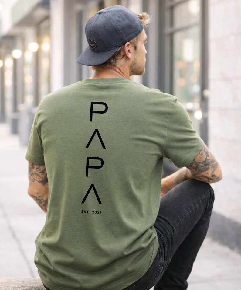 May include: Olive green t-shirt with the word "PAPA" printed vertically in black, with "EST. 2021" below. The shirt is worn by a person wearing a dark gray baseball cap and black jeans.
