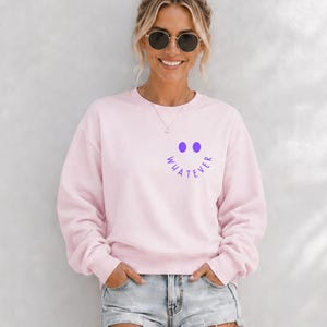 Könnte beinhalten: Hellrosa Sweatshirt mit dem Wort "WHATEVER" in Lila, angeordnet in einem Halbkreis über einem Smiley-Gesicht. Das Sweatshirt hat lange Ärmel und einen Rundhalsausschnitt. Das Model trägt Jeansshorts und eine Sonnenbrille.