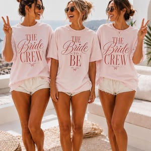 May include: Three light pink t-shirts with dark red text. Two shirts feature the text 'THE Bride CREW EST. 2026' with a small bow graphic. The center shirt features the text 'THE Bride TO BE EST. 2026' with a small bow graphic. All shirts have short sleeves.