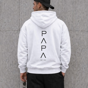 May include: White hoodie with the word "PAPA" in black vertical text on the back, with "EST. 2021" below. The hoodie has a hood and long sleeves. The person in the image is wearing a black baseball cap and black pants.