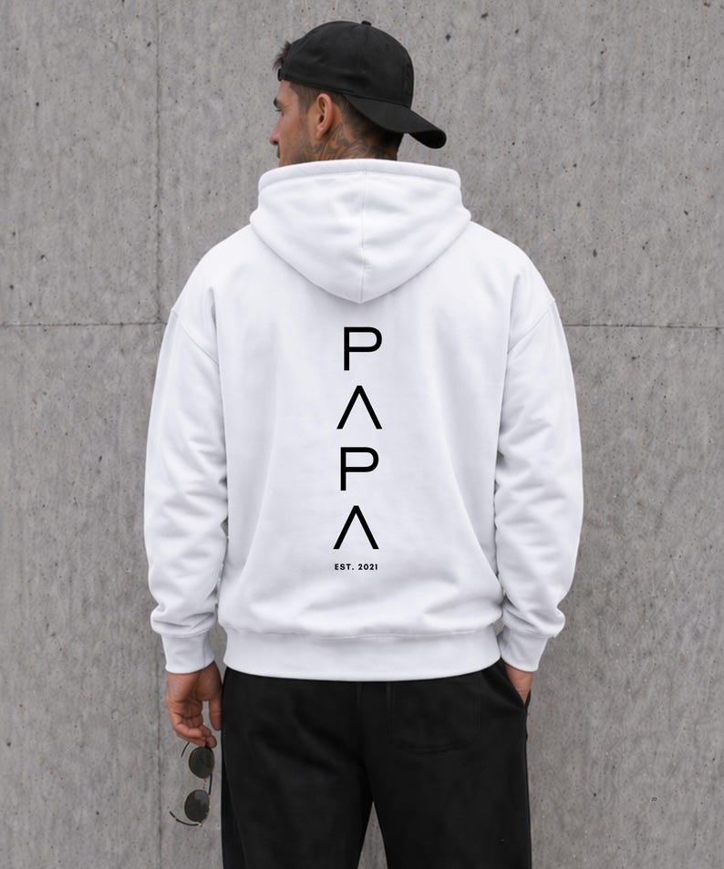 May include: White hoodie with the word "PAPA" in black vertical text on the back, with "EST. 2021" below. The hoodie has a hood and long sleeves. The person in the image is wearing a black baseball cap and black pants.