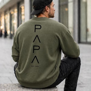 May include: An olive green sweatshirt with the word "PAPA" printed vertically in black on the back, along with "EST. 2021". The person in the image is wearing a black baseball cap and black jeans.
