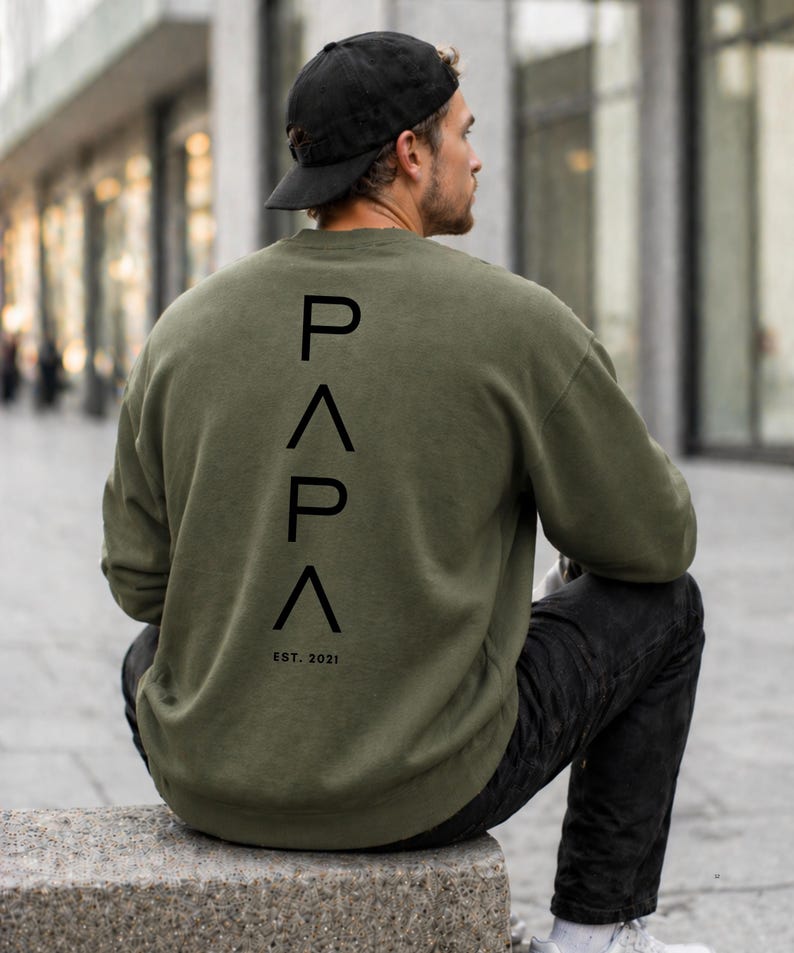 May include: An olive green sweatshirt with the word "PAPA" printed vertically in black on the back, along with "EST. 2021". The person in the image is wearing a black baseball cap and black jeans.