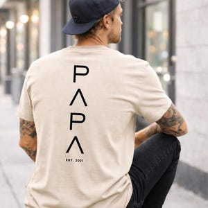 May include: Beige t-shirt with the word "PAPA" printed vertically in black capital letters, with "EST. 2021" below. The shirt is worn by a person wearing a navy blue baseball cap and black jeans.