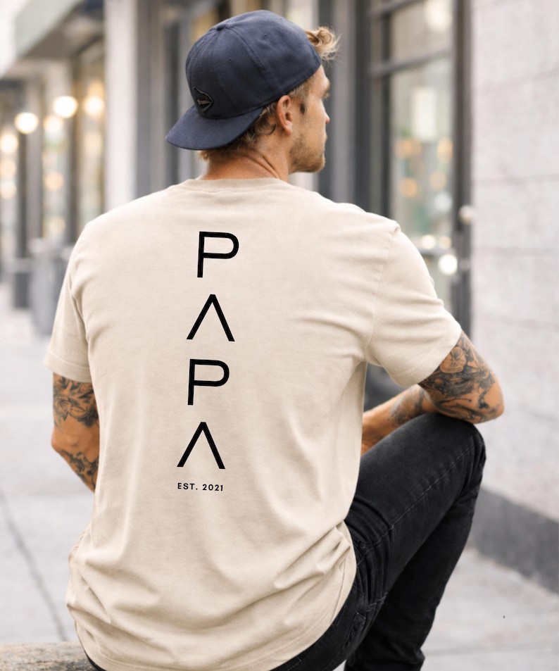 May include: Beige t-shirt with the word "PAPA" printed vertically in black capital letters, with "EST. 2021" below. The shirt is worn by a person wearing a navy blue baseball cap and black jeans.