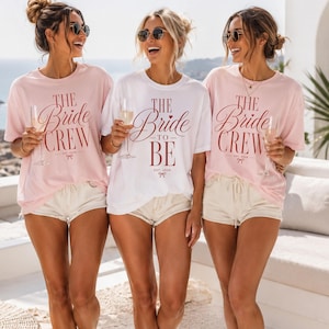 May include: Two light pink t-shirts with the text THE BRIDE CREW EST. 2026 and a white t-shirt with the text THE BRIDE TO BE EST. 2026 in red lettering. Each shirt features a small red bow graphic below the text and short sleeves.
