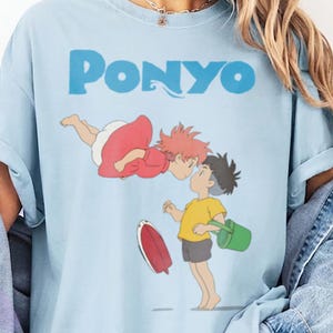 May include: A light blue t-shirt featuring a graphic print of the characters Ponyo and Sosuke from the film Ponyo. The text PONYO is printed in blue above the characters. The shirt has short sleeves and a crew neckline, worn with a denim jacket.