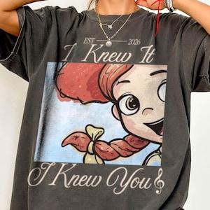 May include: A charcoal gray short-sleeve t-shirt featuring a graphic print of a cartoon character with red hair in braids. The text reads "EST 2026", "I Knew It", and "I Knew You" with a musical note symbol. The shirt has a relaxed, loose fit.