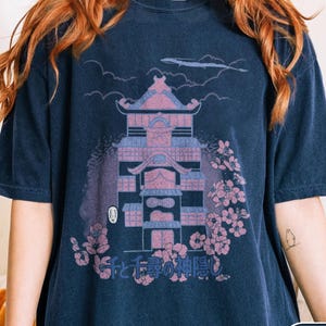 May include: A navy blue short-sleeve t-shirt featuring a graphic print of a multi-story building with pink and blue accents, surrounded by cherry blossoms and clouds. The Japanese text "千と千尋の神隠し" is printed below the building illustration.