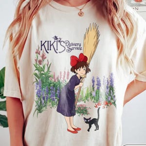 May include: An ivory short-sleeve t-shirt featuring a graphic of Kiki from Kiki's Delivery Service holding a broomstick, accompanied by a black cat. The design includes purple and green flowers and the text Kiki's Delivery Service in dark blue.