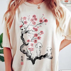 May include: An ivory short-sleeve t-shirt featuring a graphic print of a dark, stylized tree with pink cherry blossoms. Three small, white, round-headed figures are positioned on the branches. Black Japanese characters are printed vertically.