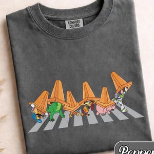 May include: Pepper-colored t-shirt featuring a graphic of Toy Story characters crossing a crosswalk, each wearing an orange traffic cone. The shirt is made by Comfort Colors.