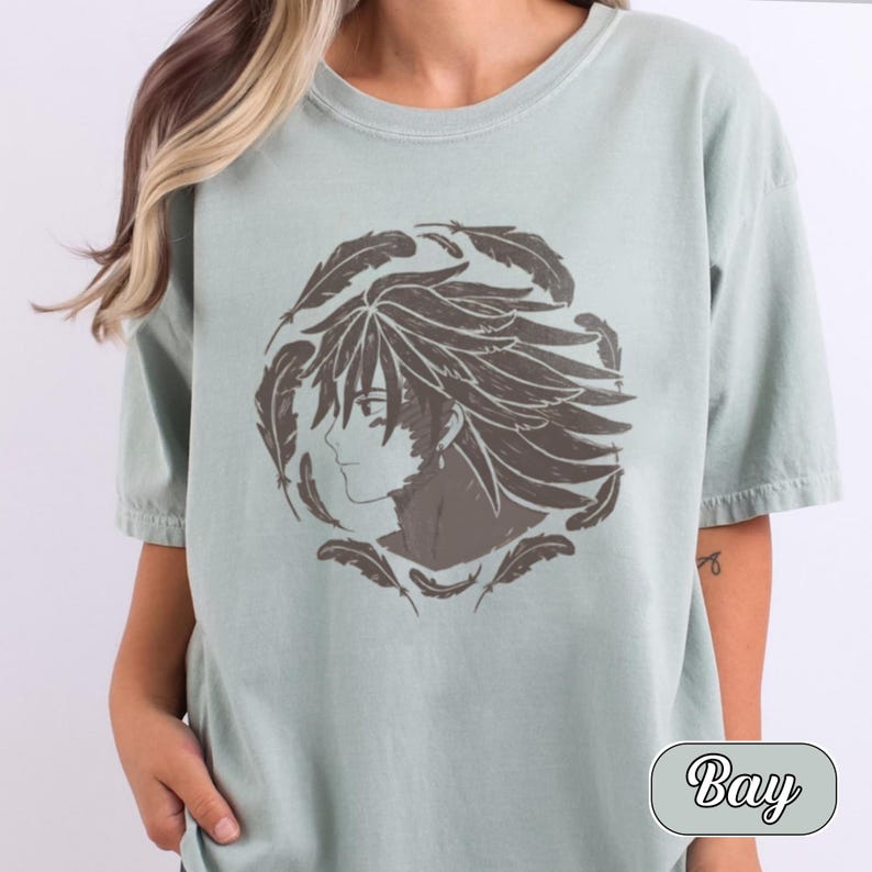 Pu&ograve; includere: Una t-shirt a maniche corte color verde salvia chiaro con una stampa grafica circolare marrone scuro. Il design raffigura il profilo di un personaggio in stile anime con capelli a punta circondato da un anello di piume fluttuanti. La parola Bay &egrave; stampata.